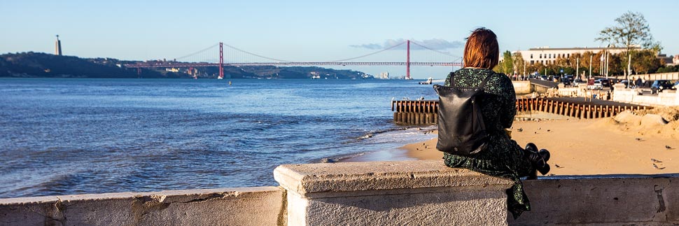 Ein Frau sitzt am Stadtstrand von Lissabon mit Blick auf die Brücke des 25. April
