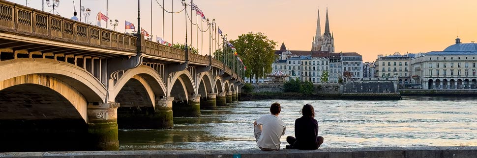 Ein Paar sitz am Ufer der Adour und blickt im Sonnenuntergang auf die Altstadt und die Pont Saint-Esprit in Bayonne, Frankreich