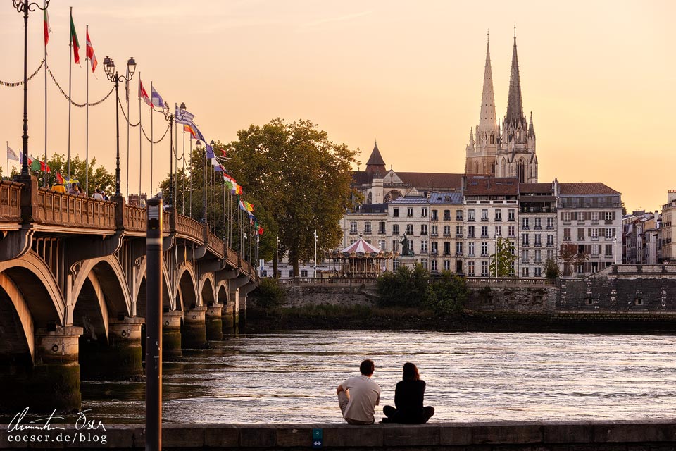 Ein Paar sitzt im Sonnenuntergang am Ufer der Adour und blickt auf die Pont Saint-Esprit und die Altstadt von Bayonne mit der Kathedrale