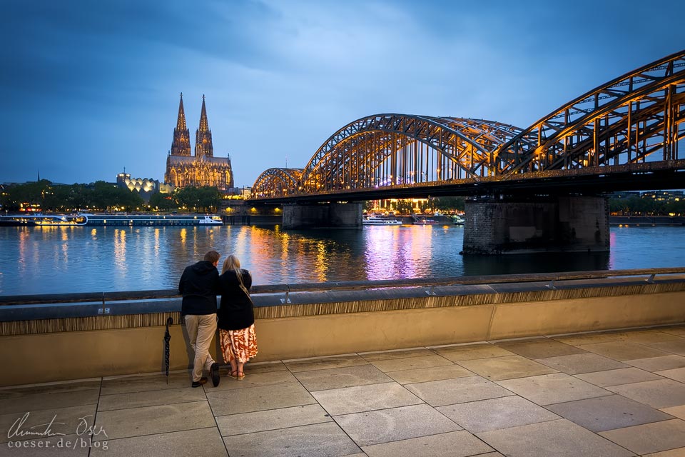 Paar blickt in der blauen Stunde vom Aussichtspunkt Rheinboulevard auf den Kölner Dom und die Hohenzollernbrücke