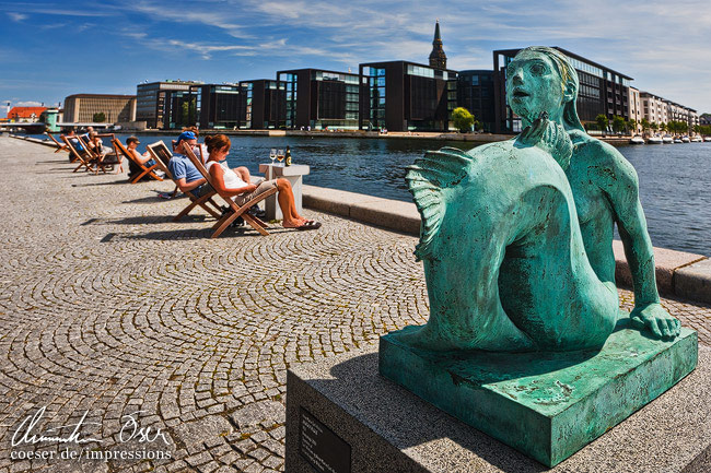 Eine Skulptur neben Menschen, die die Sonne genießen vor der 'Black Diamond'-Bibliothek in Kopenhagen, Dänemark.